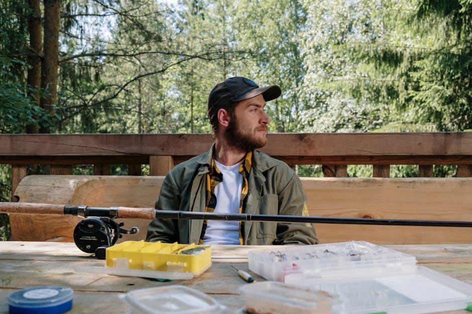 Man with beard sitting outdoors with fishing equipment, capturing nature's essence.